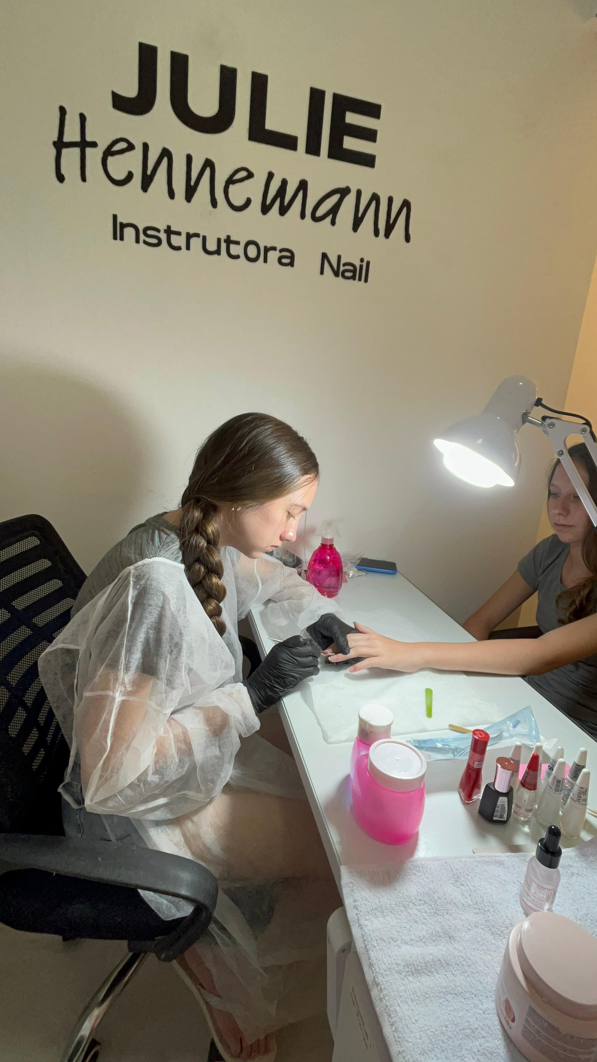 Aluna com jaleco e luvas realizando cutilagem em uma modelo, em frente ao letreiro Julie Hennemann Instrutora Nail